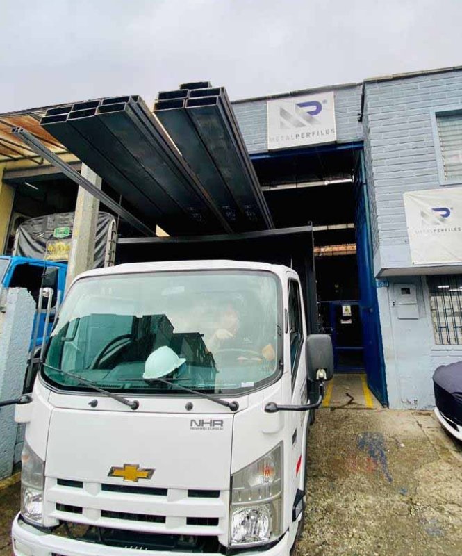 Camión blanco con tubería rectangular cargada frente a una bodega gris
